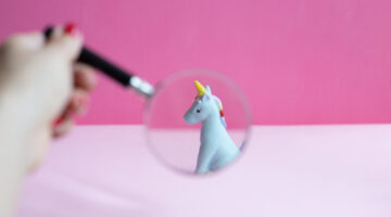 A hand holding a magnifying glass over a blue unicorn on pink background.